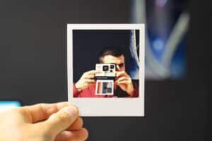 Hand holding a Polaroid photo of person in red shirt aiming a Polaroid camera, creating a layered visual against dark background.