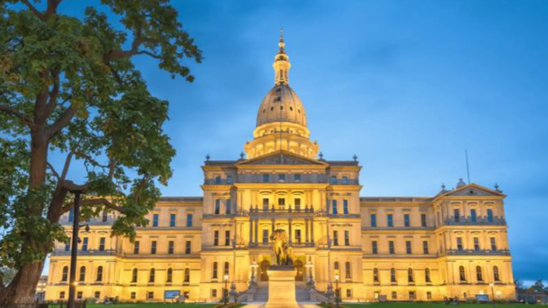 Michigan State Capitol in Lansing lit at dusk, featuring its domed architecture, front statue, and surrounding trees.
