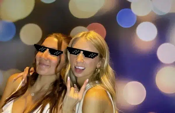 Two guests in white outfits posing with pixelated “deal with it” sunglasses, peace and rock hand gestures, colorful bokeh light backdrop