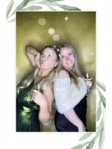 Two guests in formal dresses holding drinks at a FastBooth photo booth with golden backdrop and leafy border