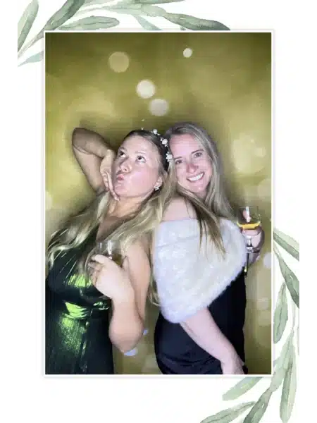 Two guests in formal dresses holding drinks at a FastBooth photo booth with golden backdrop and leafy border