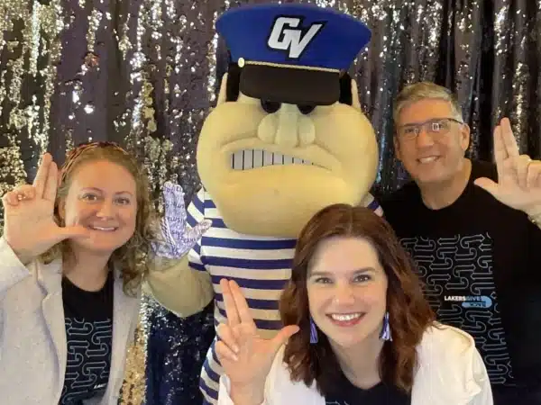 Three guests smiling with Louie the Laker mascot in blue striped shirt and GV hat, sequined backdrop, showing “L” hand gesture for school spirit
