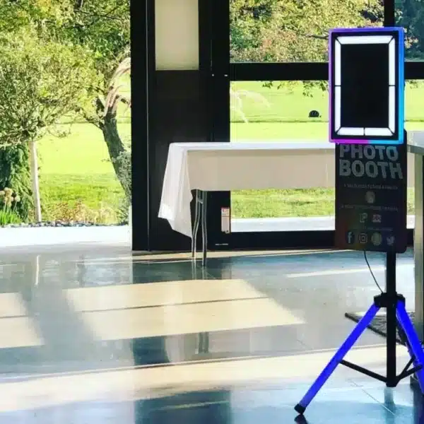 Modern LED tripod photo booth with illuminated frame and Photo Booth sign set up at indoor event space near window and table
