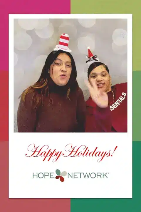 Two people in festive hats posing before holiday background with Happy Holidays text and Hope Network logo